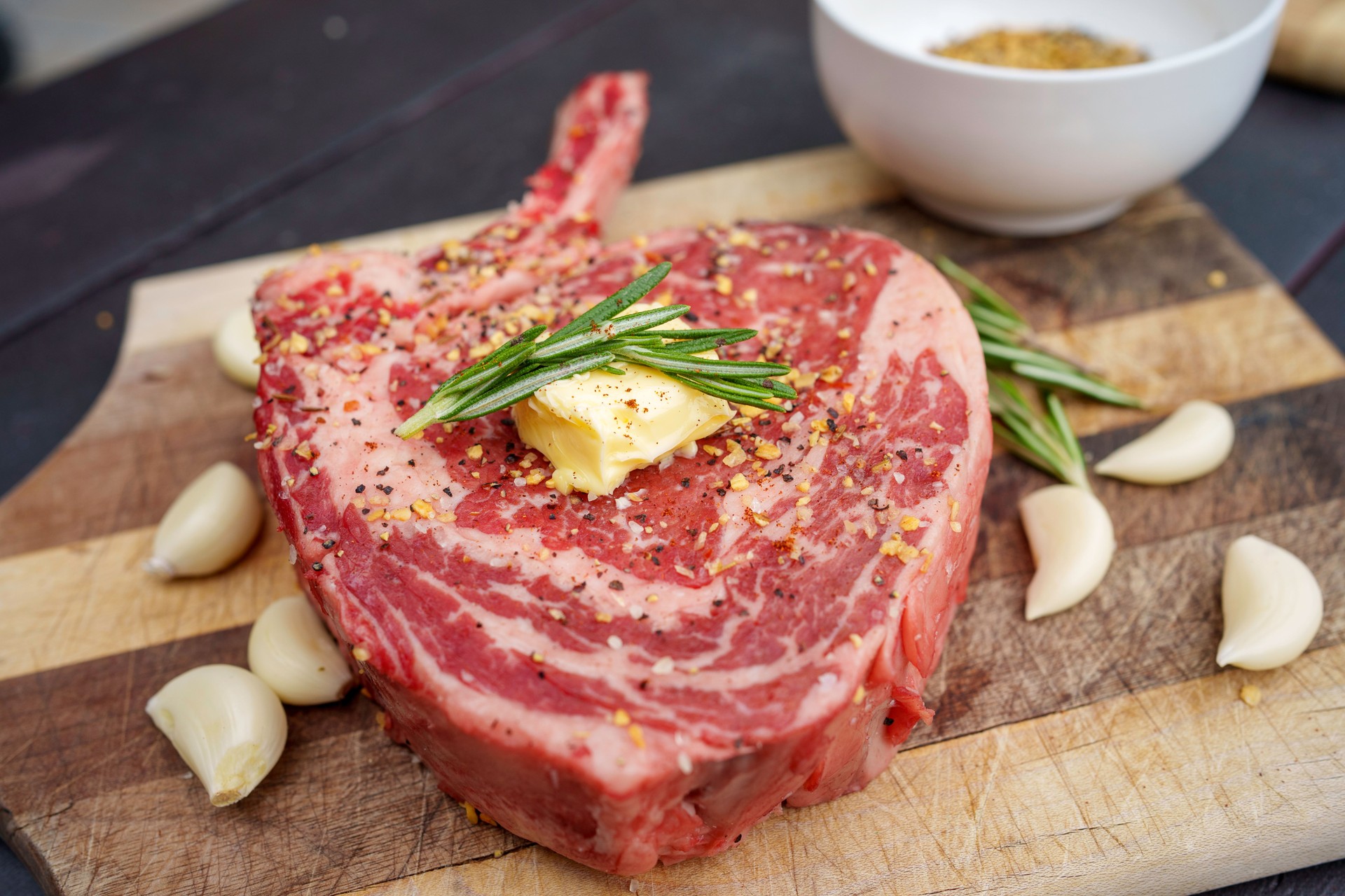 Raw Tomahawk Rib Eye Beef Steak on a Rustic Wooden Cutting Board Ready to Cook Next to a Charcoal Grill in a Residential Back Yard in Summertime With Garlic Cloves and Rosemary